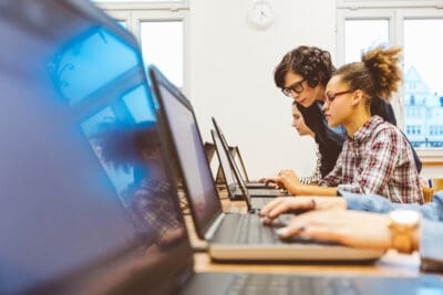 Several people work on laptops in a classroom setting. One person stands, assisting another seated person.