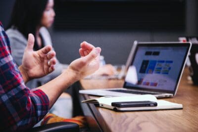 Person gesturing with hands during a meeting at a table, with a laptop displaying charts and a notebook and phone nearby. Another person is in the background.