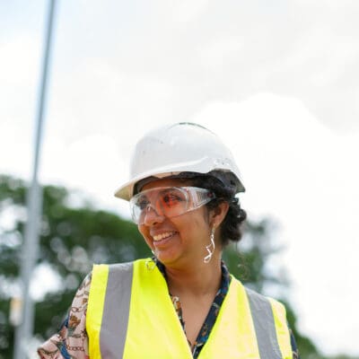 Person smiling, wearing a white hard hat, safety goggles, and a yellow reflective vest, standing outdoors with blurred greenery in the background.