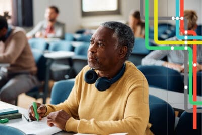 An older man sits attentively in a classroom, wearing a yellow sweater and headphones around his neck. Several other people are seated in the background.