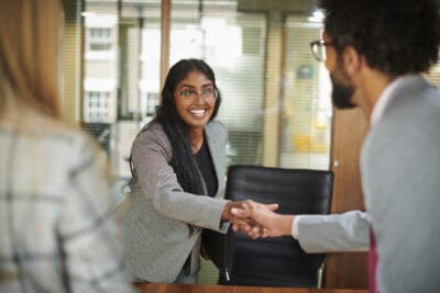 Two people shaking hands in an office setting. The woman is smiling, wearing glasses, and a gray blazer. The man's back is turned, wearing a light-colored suit. Another person's shoulder is visible.