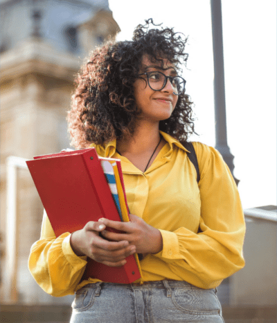 Person with curly hair and glasses in a yellow shirt, holding red folders and books, stands outside near a large building.