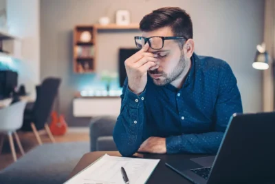 Man sitting at a desk with a laptop and paper, pinching the bridge of his nose in apparent frustration or fatigue.