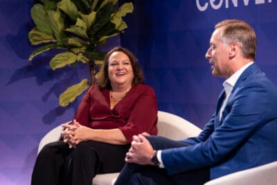 Two people seated on white chairs engaged in conversation on stage with a potted plant in the background.