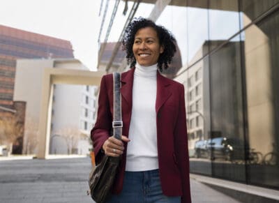 A person in a burgundy blazer and white turtleneck walks outside by modern buildings, carrying a shoulder bag.