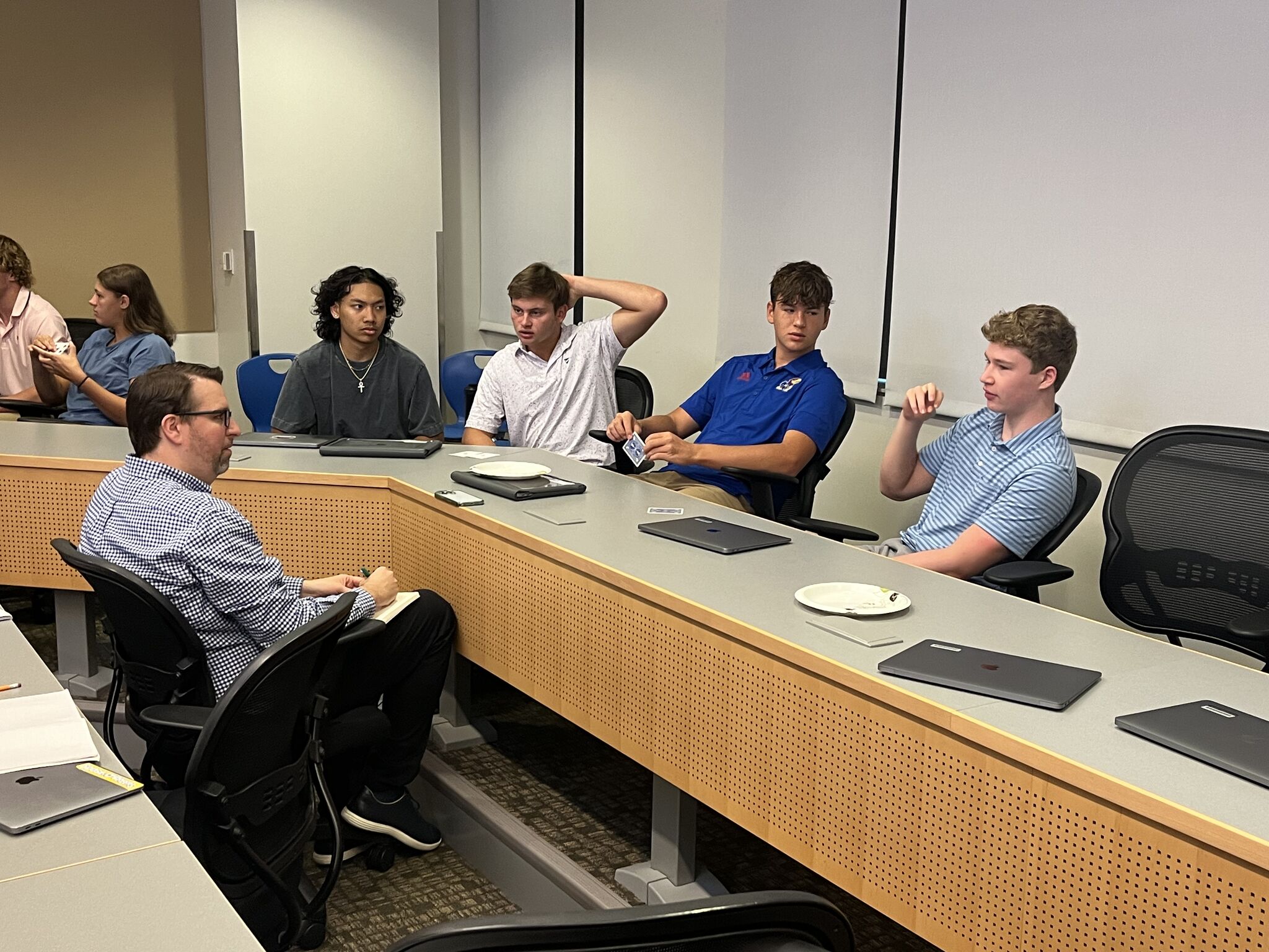 A group of people sitting at a conference table, engaged in discussion. Laptops are placed on the table.