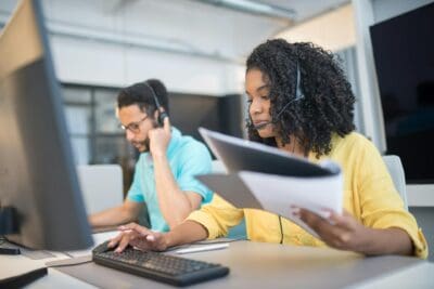 Two people wearing headsets work at desks with computers. The woman in the foreground holds documents and types, while the man in the background talks on the phone.