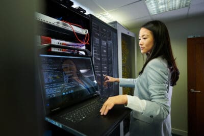 A woman in a server room works on a laptop connected to a server rack, surrounded by cables and equipment.