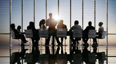 Silhouetted people in a boardroom meeting with one person standing, against a backdrop of large windows and sunset.