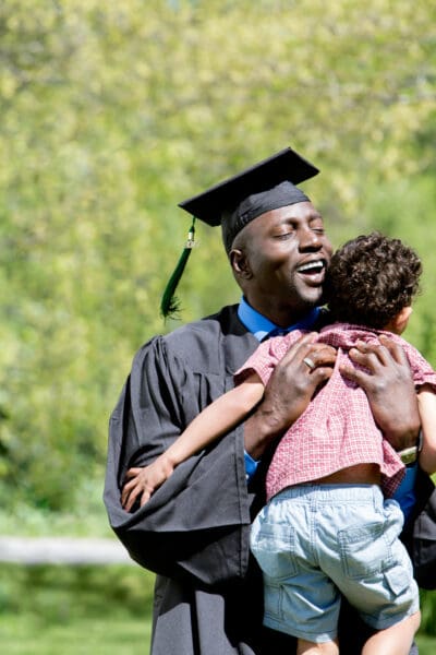 A graduate in a cap and gown joyfully hugs a child outdoors, with greenery in the background.