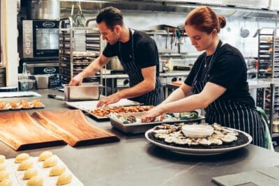 Two chefs in black aprons prepare food in an industrial kitchen, with various dishes and ingredients spread on the counter.