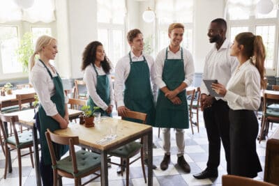 A group of restaurant staff in green aprons stands together in a dining area, listening to a woman holding a tablet. Tables and chairs fill the space, with sunlight streaming in.