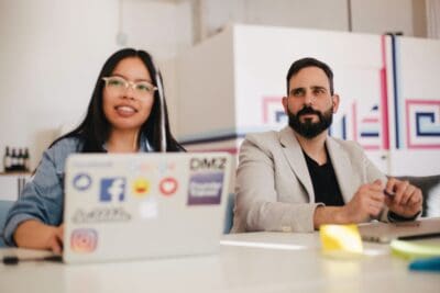 Two people sitting at a table with laptops covered in stickers. One person is smiling; the other appears attentive. They are in a bright room with modern decor.