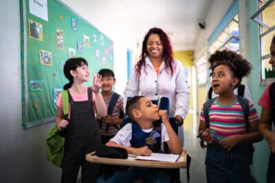 A group of children walk down a school hallway with a teacher. One child is in a wheelchair, accompanied by the teacher, while others chat and smile. A bulletin board is visible on the wall.