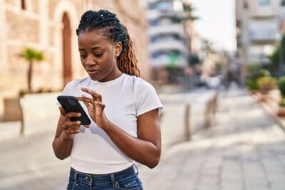 A person stands on a cobblestone street, focused on using a smartphone. They wear a white t-shirt and jeans. Buildings and palm trees are visible in the background.