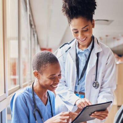 Two medical professionals, one in a white coat and the other in scrubs, smiling and looking at a tablet near a window in a hospital corridor.