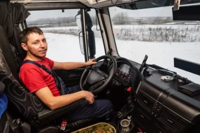 A man in a red shirt and overalls sits inside a truck cabin with snow outside. He's holding the steering wheel and looking at the camera.