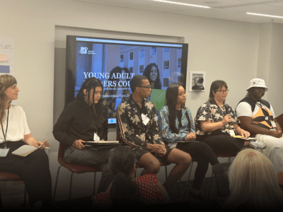 Panel of six individuals seated, participating in a discussion. A presentation slide titled 