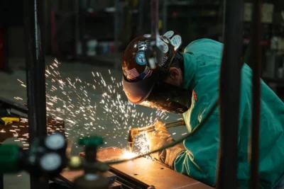 A welder in green protective clothing and a helmet works on metal, creating sparks in a workshop setting.