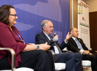 Three individuals seated on a panel discussion. The man in the middle is speaking while holding a microphone. A blue backdrop and a banner with text are behind them.