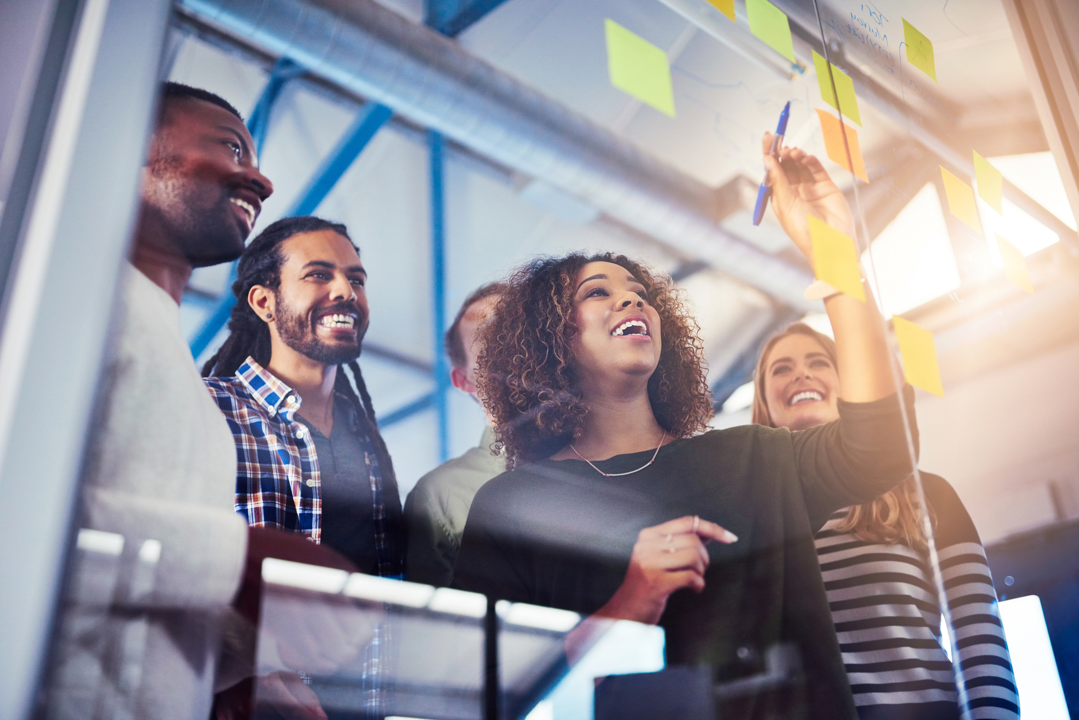 A group of people smiling and collaborating, with one person writing on sticky notes placed on a glass wall.