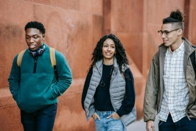 Three people walking and smiling, one wearing headphones, against a red brick wall.