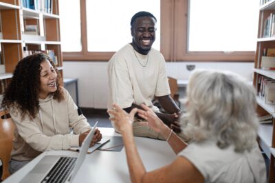 Three people sitting around a table in a library, laughing and talking. A laptop is open on the table.