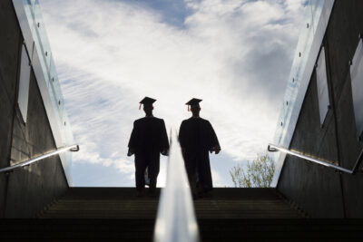 Two graduates in caps and gowns walk up a concrete stairway, silhouetted against a bright sky.