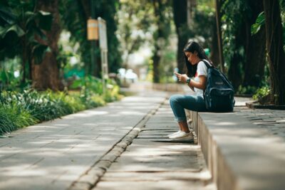 A person sits on a low wall along a tree-lined path, looking at their phone. They have a backpack and are dressed casually in jeans and a t-shirt.