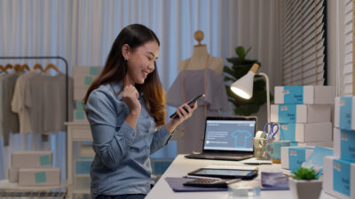 A woman in a blue shirt smiles at her phone in a home office, surrounded by packages, a laptop, and a tablet on a desk.