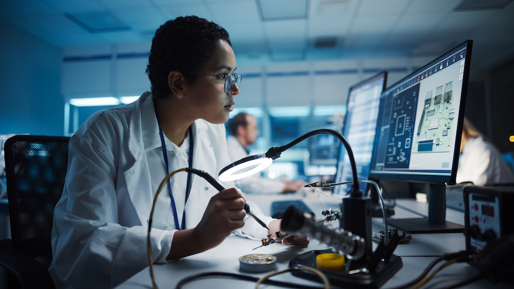 A person in a lab coat uses a magnifying lamp to examine electronics on a circuit board, with a computer displaying technical diagrams in the background.